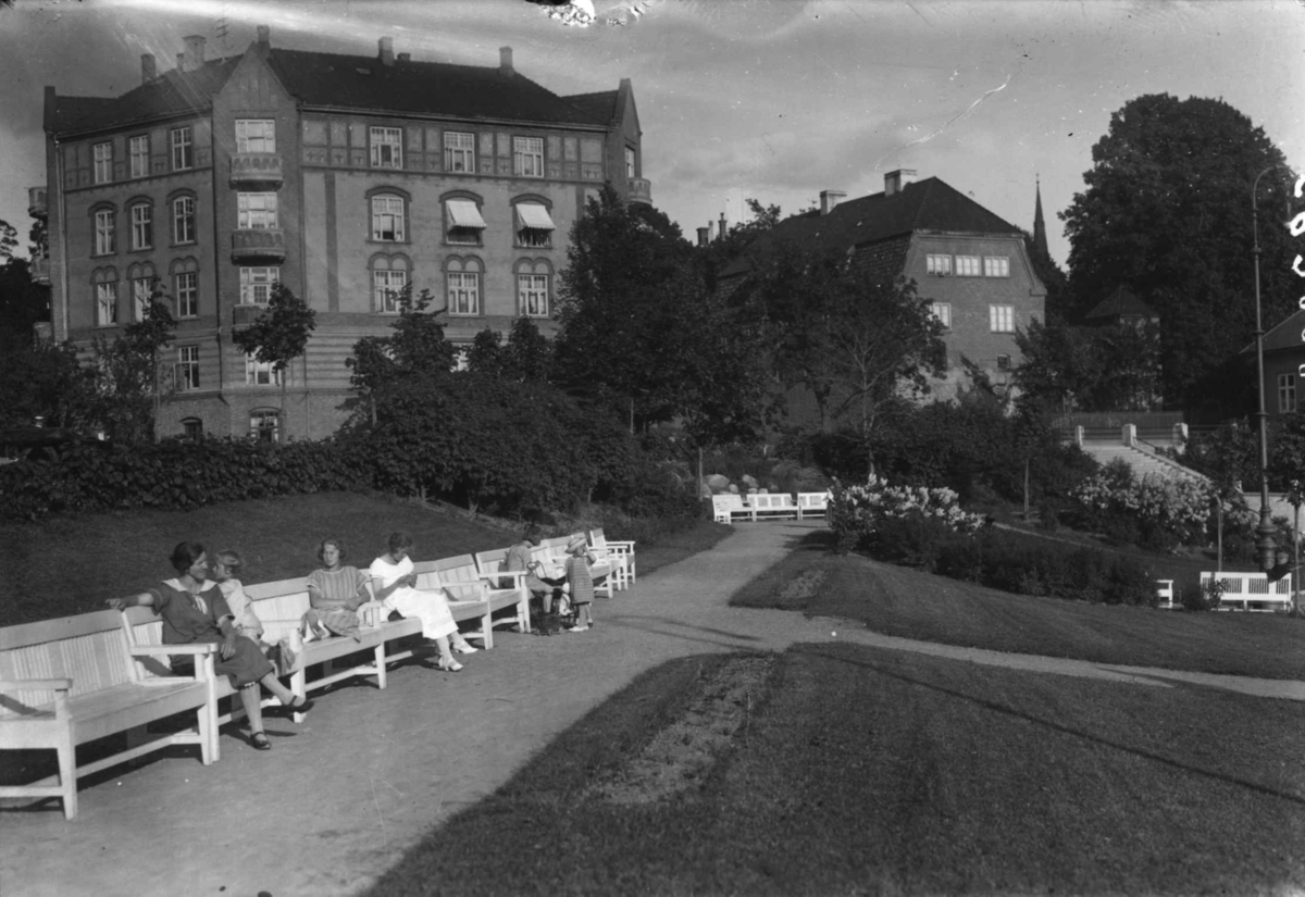 Oslo. Mennesker sitter på benker i en park ved Vestkanttorvet.  Bybebyggelse i bakgrunnen. Trær. 