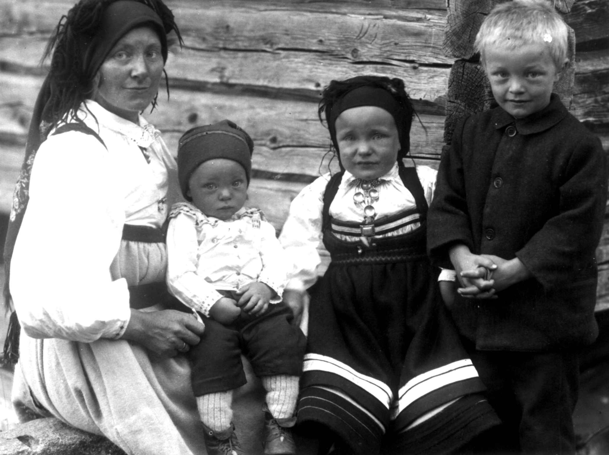 Kvinne- og barnedrakt, gruppeportrett, Valle, Setesdal, Aust-Agder, antatt 1924. Kvinne med tre barn sittende utendørs foran tømmervegg. Fra "De Schreinerske samlinger" (skal oppgis).