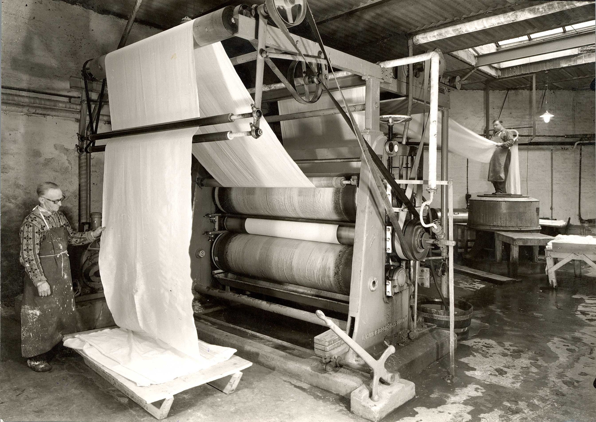 Produksjon ved A/S Joh. Petersens Linvarefabrik. Tekstil går gjennom maskin med store valser. To menn arbeider i hver sin ende.