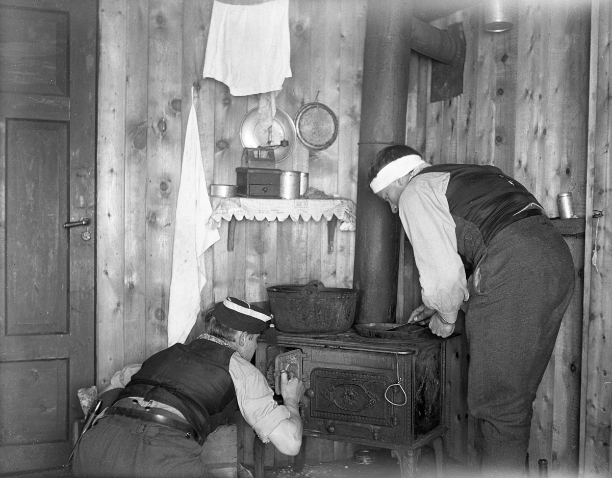 To menn står for vedfyring og matlaging på hytte. Fotografert påsken 1923.