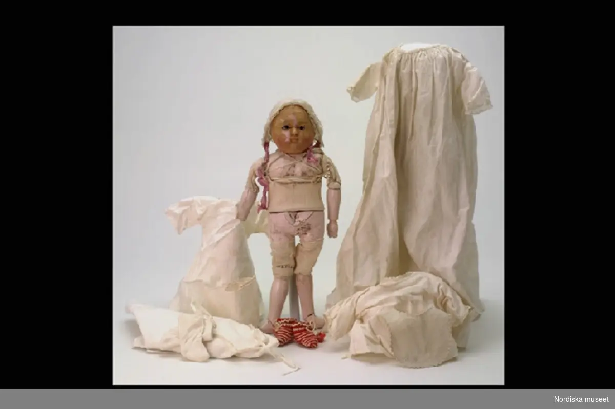 Inventering Sesam 1996-1999:
H 44 (cm)
Docka, baby, lindebarn, sk "Täufling" (a), med kläder (b-h), kropp av Motschmann-/ Lindner-typ, med vridbart huvud, bröstparti, bäcken, händer och fötter av massa, underarmar och underben av trä, rosa bemålning, och överarmar, mellangärde och lår klädda med vit textil. Ansiktet överdraget med vax (krackelerat), målade ansiktsdrag, svarta blundögon av glas och målade babylockar. Ljudbälg i magen (defekt).
Iklädd linne (b), tröja (c), blöja (d), linda (e), strumpor (f:1-2), virkade sockor av vit- och rödrandigt ullgarn, knytband med tofsar (g:1-2) bärklänning (h)  haklapp (i), mössa med knytband av violett siden (j) och mössa, glest virkad av lingarn (k). Tillhörande tröja, inv nr 156.551.
"Motschmann" och "Lindner", se Coleman, Dorothy S, "The Collector's Encyclopedia of doll's", vol. II, New York 1986.
Jfr inv nr 107.728, 113.407 och 252.577.
I dåligt skick; vaxskiktet krackelerat, bäckenpartiet skadat, huvudet löst, en hand saknas.
Anna Womack februari 1997