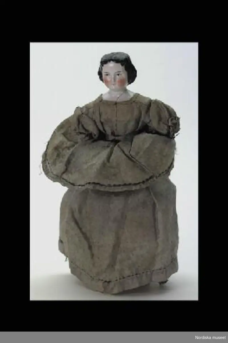 Katalogkort:
"Docka, 1860-tal. L 25 cm. Huvud och ett ben (resten saknas) porslin, stoppad kropp, klänning blått mönstrat siden, 2 underkjolar och byxor av vit bomullslärft."