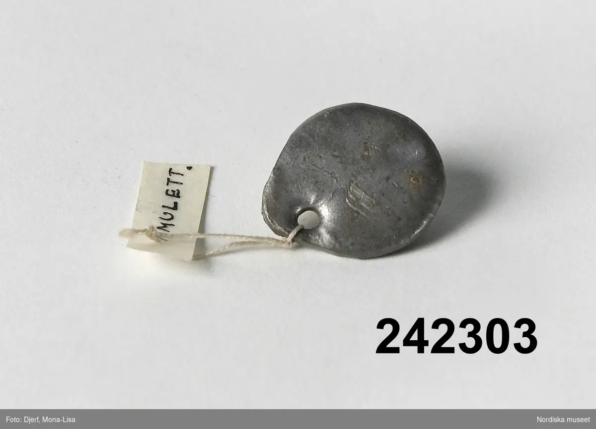 Huvudliggaren: 
"Amulett, av bly (eller tenn), gjuten bricka av litet format och liknande ett hängsmycke; litet hål för snöre eller kedja. Använd mot 'skorven' (Engelska sjukan). Ink. 13/3 1951 av Fru Swea Johansson, Urshult."

Bilaga:
"2 amuletter  [avser inventarienr 242 302 och 242 303] , olika som tros varit på ett band om halsen, mot engelska sjukan. Vidskepelse av denna typ var på 1800-talet vanlig. Metall skrapades av och åts även av kreatur. Englanders Tingsryd."