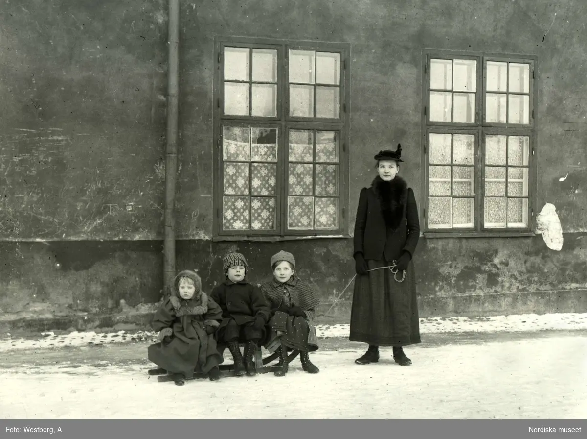 Kvinna med tre barn på en kälke framför hus.