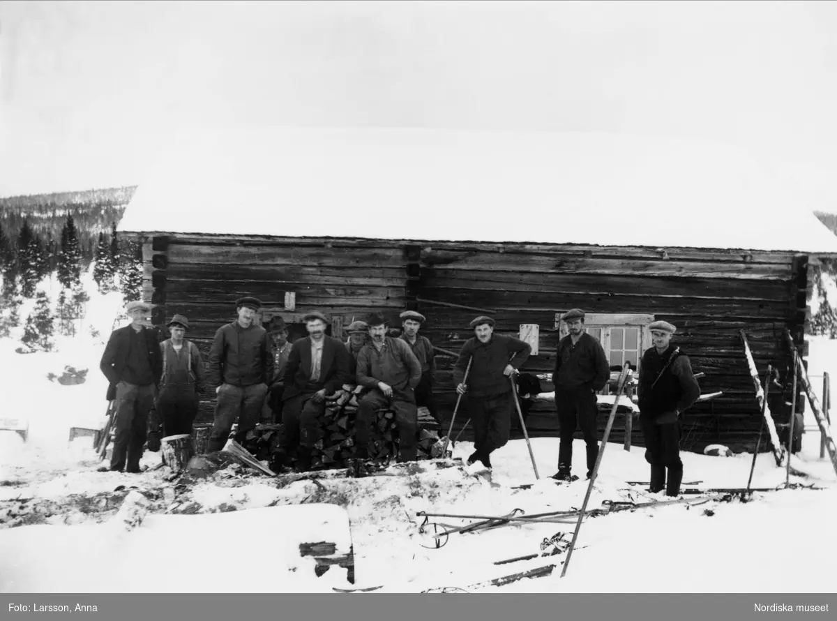 Gruppbild. Elva män, stående utanför timmerhus i Långsätern Värmland.