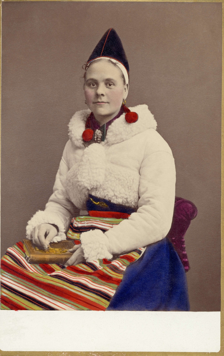 Porträtt av Täpp Margit Ersdotter iklädd folkdräkt från Rättvik i Dalarna med vinterjacka. I knät håller hon en bok. Kolorerat fotografi.