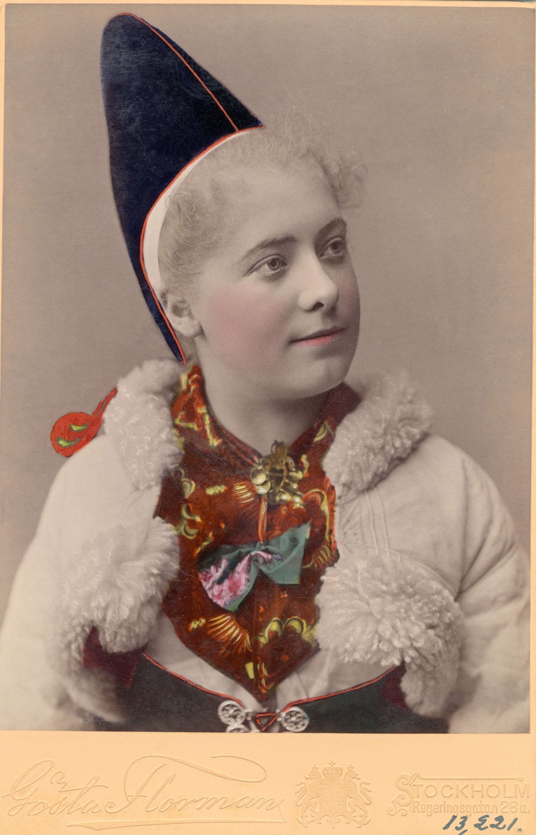 Bröstbild av en ung kvinna i folkdräkt, från Rättviks socken i Dalarna, med en pälsbesatt vit jacka. Kolorerat fotografi.