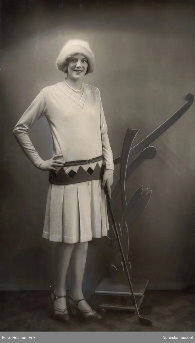 Kvinna iklädd stickad kjol med tillhörande överdel, tvåfärgade skor, huvudbonad och en golfklubba i handen. April 1929. Nordiska Kompaniet.