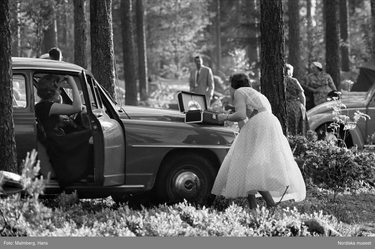 Till dans. Midsommar i Leksand, Dalarna 1955. Kvinnor sminkar sig vid bil i skogsområde .