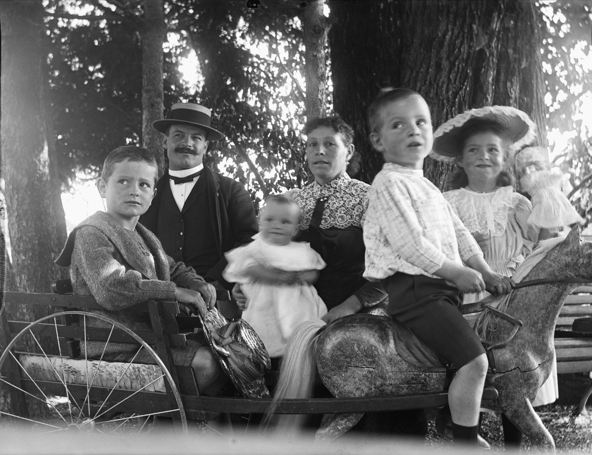 Gruppbild, familjebild. Man och kvinna med fyra barn poserar runt leksakshäst med vagn i park.