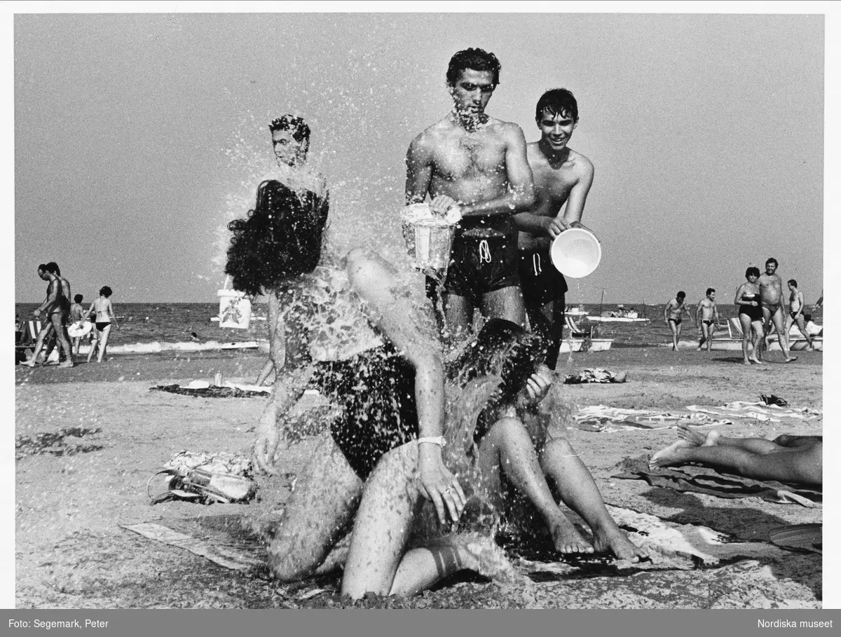 Strandliv. Lek och stoj på en solig sandstrand där tre unga män i badbyxor slänger vatten på två unga kvinnor som sitter på ett badlakan. De använder små leksakshinkar av plast. I bakgrunden solande, badande och vandrande människor, havet och några små båtar.