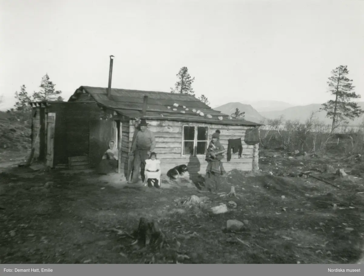 Johan Turi står vid Gruvstugan, tillsammans med en arbetare och hans fru och dotter. Kläder hänger till tork, en hund ligger på marken.