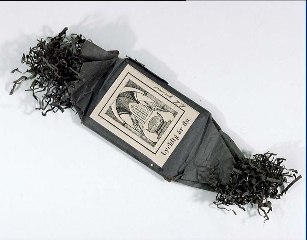 Begravningskaramell, svart silkepapper. Svartvitt märke med likkistmotiv med text: "Lycklig du är".