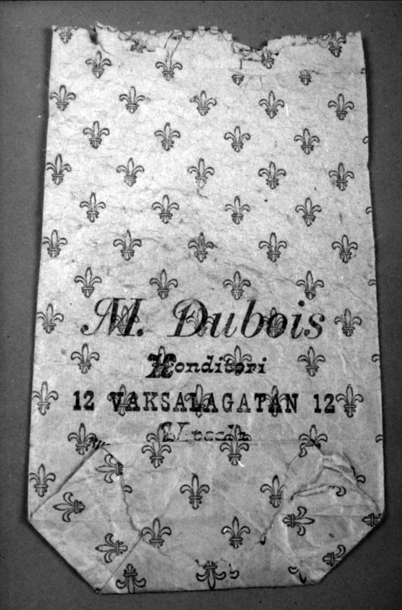 Påse av vitt papper med blått mönster i form av franska liljor och blå text: M.Dubois, Konditori, Vaksalagatan 12, Upsala.