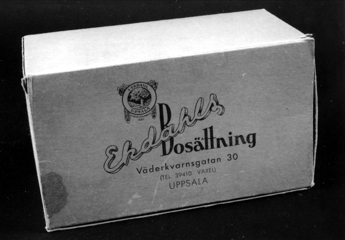 Kartong av gråblå blekt papp, hopfogad med metallklamrar, fällock med firmamärke och text i blått: Ekdahls Bosättning, Väderkvarnsgatan 30, Uppsala.