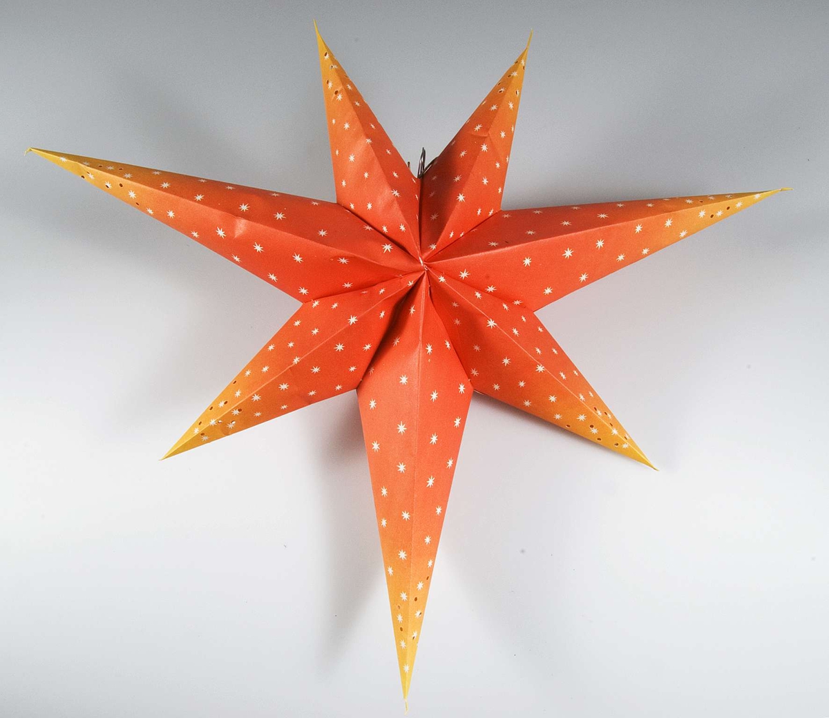 Adventsstjärna av papper med elektrisk lampa och sladd. Stjärnan rödorange med vita stjärnor och perforerad i ändarna.
