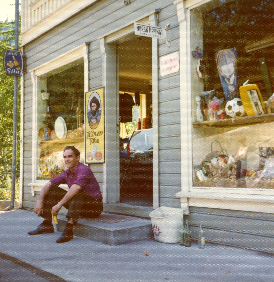 En mann sitter utenfor en butikk og røyker en sigarett.