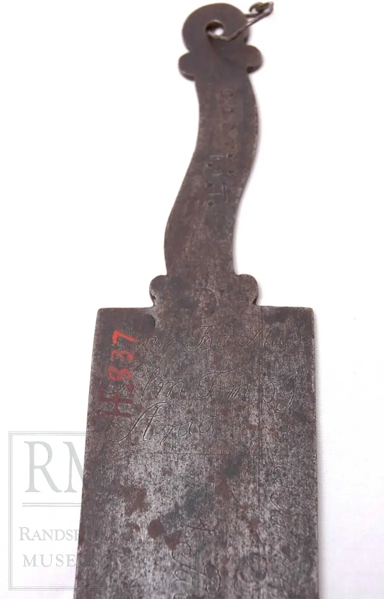 Rektangulær plate av jern med håndtak i ene enden. Håndtaket har en liten bøy. Merker viser 1-12 tommer