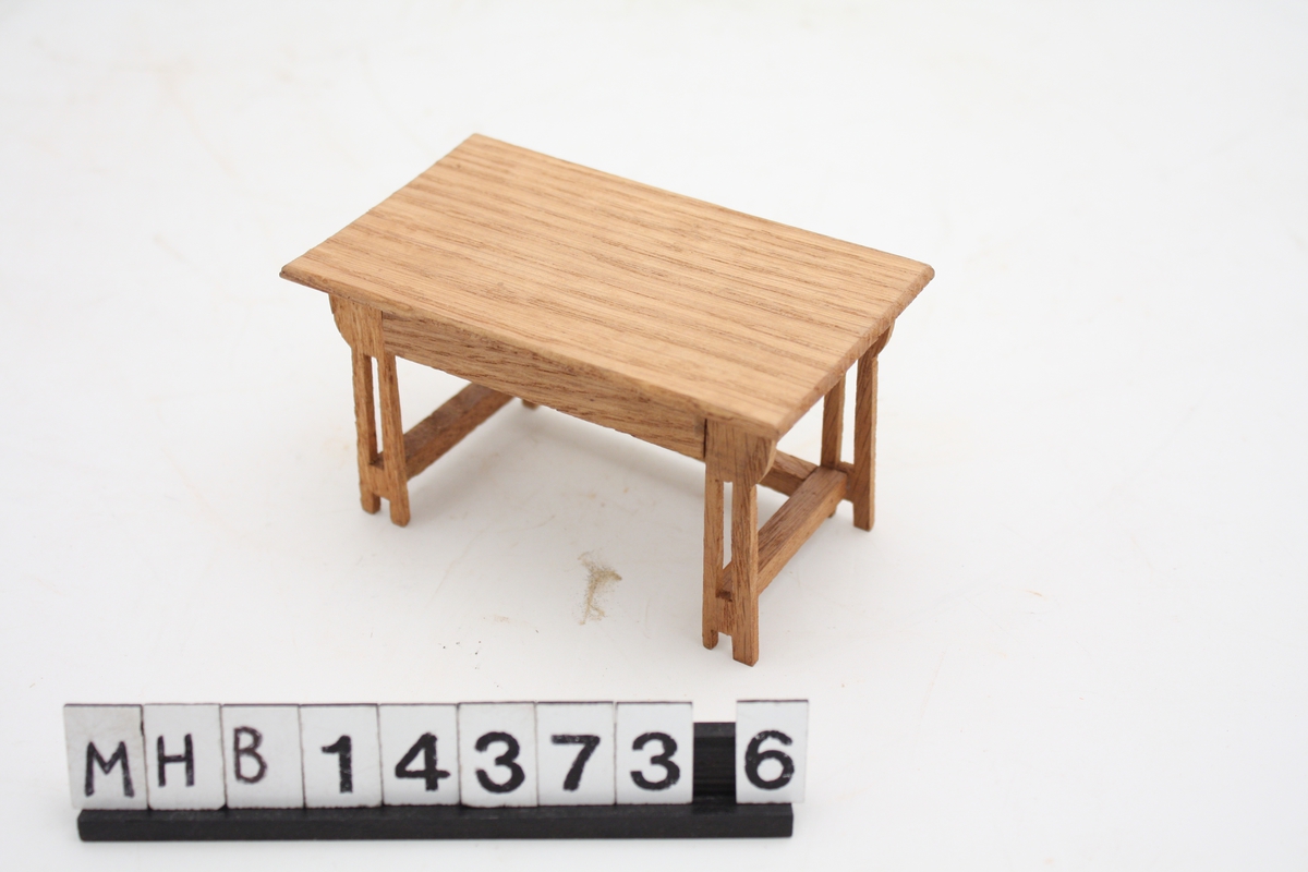 Miniatyrbord til dukkehus. Det er i tre, og har bordplate med rektangulær form. I hvert hjørne er doble bordbein som nede er forbundet med hverandre både i lengde- og bredderetning.