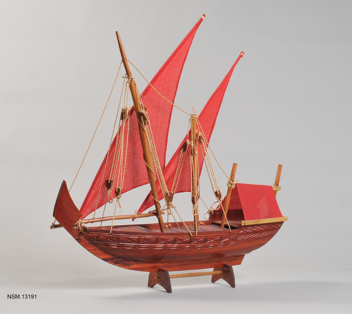 Arabisk dhow. Enkel souvenirmodell, lyst brunt treverk med utskjæringer i fribord og på dekk, to master med røde seil, rød presenning over akterdekket.