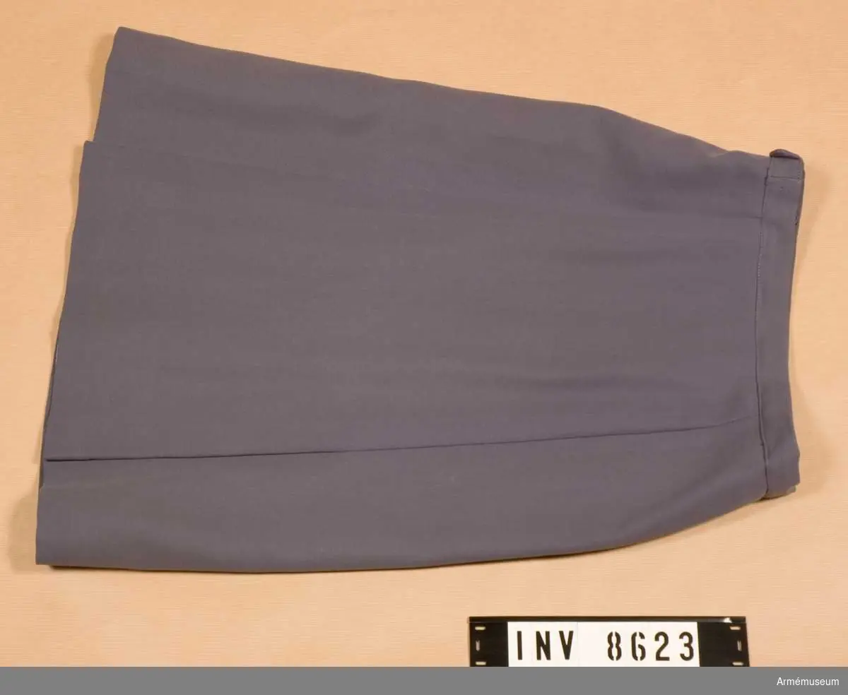 Samhörande gåva 8617-8626.
En blågrå något utställd kjol med linning, blixtlås i vänster  sida och veck på var sida av den infällda framvåden. Kjolen är fodrad med grått siden. Den är både nedlagd och utsläppt i sömmarna bak vilket syns i de ursprungliga pressvecken. Hettermarks konfektionsfirma. Källa SLK Värt att veta... 1970 sid 18.