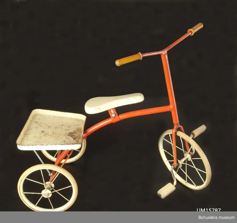 Trehjulig barncykel med röd stomme, vit sadel och pakethållare. Gummiklädda handtag, trampor av hårdgummi.