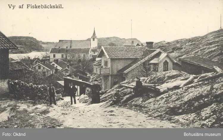 Vägen Repelia i Fiskebäckskil, tre personer står och tittar in i kameran, en sitter längs vägen. I bakgrunden syns hustak och Fiskebäckskils kyrka.