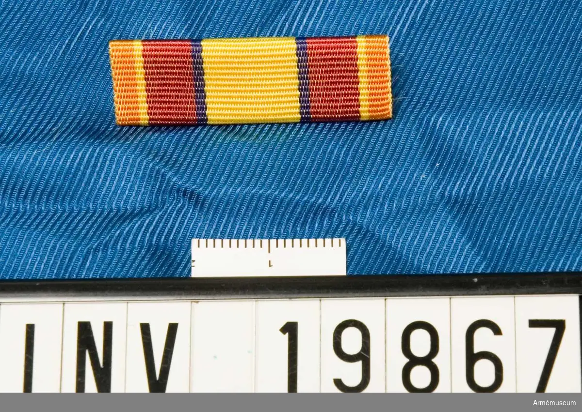 Bandet är mörkrött med en bred gul rand på mitten på vardera sidan åtföljd av ett blått streck samt med orange kanter åtföljda av ett gult streck. Släpspännet förvaras i ask tillsammans med en medalj och en miniatyrmedalj.