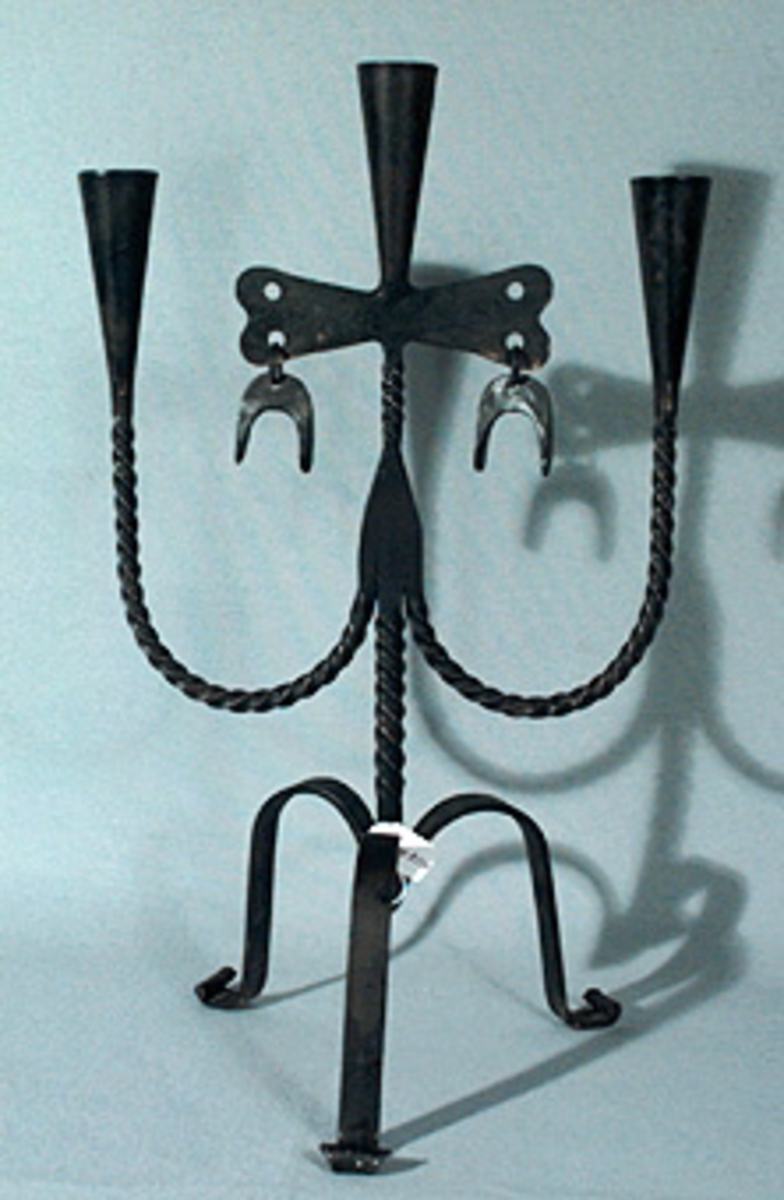 Trebent ljusstake, benen i bandjärn. Tre ljushållare, armarna spiralvridna. Ljushållaren i mitten har en figur med hästskoliknande hängen på var sida om armen.