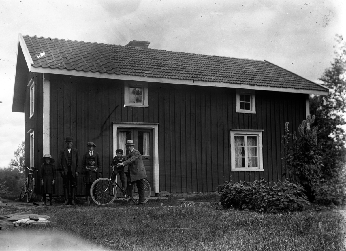 Två män med cyklar och fyra barn står framför ett tvåvånings trähus.