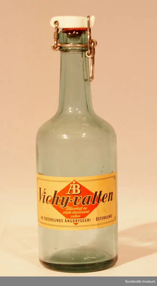 Flaska i gröntonat glas med patentkork från AB  Östersunds Ångbryggeri. Etikett med texten 'ÅB Vichy-vatten, Tillverkat av ozon-steriliserat vatten, AB ÖSTERSUNDS ÅNGBRYGGERI, ÖSTERSUND'.