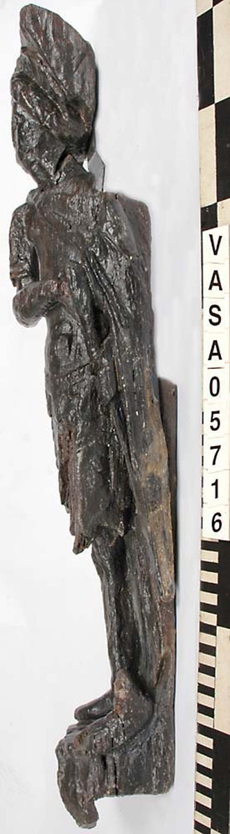 Skulptur föreställande mansfigur iklädd romersk rustning. Huvudet pryds av en lagerkrans som avtecknar sig mot ett stort snäckskal. Ansiktet med den rynkade pannan är slätrakat. Figuren är iförd thoraxrustning med en utmed ryggen nedhängande mantel. Manteln är draperad i en smal sträng snett över bröstet, och sitter fästad med ett spänne på höger skuldra.



Figurens huvud och överkropp är helt vridna mot vänster, underkroppen endast till hälften. Benen är frontalställda. Höger arm hålls vinkelrätt böjd över bröstet. Kroppstyngden vilar på höger ben, medan vänster ben hålls böjt. Upptill är bakstycket framdraget i en enkel, rektangulär "huvudplatta".



Figuren står på en sockel, som framtill pryds av en djurmaskaron, ett groteskt lejonansikte eller dylikt. Maskaronen har spetsiga rovdjurständer och rundade öron. På sockelns mitt finns en banderoll med en namninskription. Bokstäverna R och O är tydligt skönjbara på höger del av bandet, medan bokstäverna på bandets vänstra del är utplånade.



Skulpturens baksida är rak och uppvisar tre snedställda, olika djupt liggande fält. De tre fälten, som utgör anliggningsytor, ger skulpturen en trappstegsformad profil.



Skulpturen är mycket sliten. Figurens högra hand är kraftigt skadad. Vänster arm med vidhängande del av manteln saknas.



Text in English: Sculpture representing a male figure wearing Roman armour. The figure has a laurel-wreathed head, set against a large sea shell. The face is clean-shaven and the forehead wrinkled. The figure is wearing thorax armour, with the mantle hanging down the back and draped across the chest from the right shoulder to the left side of the waist in a narrow swathe.



The head and upper part of the body are turned wholly, the lowwer part of th ebody halfway, to the left. The legs are posed frontally. The right arm is held bent at a right angle cross the chest, its hand is badly damaged. The left arm and the part of the mantle that covers it has fallen off. The body rests on the right leg while the left is bent.

The upper part of the back extends upwards in a simple, rectangular  "head board".



The figure stands on a plinth which is decorated by a grotesque mask, reminiscent of a lion head with teeth and rounded ears. In the centre of the plinth is a broad label with a name inscription. As we view it from the front no letters can be seen on the left but the letters R and O are clearly visible to the right.



The sculpture''s back has three obliquely placed flat surfaces of different depths, that are areas of attachment. The flat surfaces make the sculpture looks like a step-ladder in profile.

The sculpture is badly worn. The figure''s right hand is badly damaged. The left arm and the part of the mantle which covers it is missing.