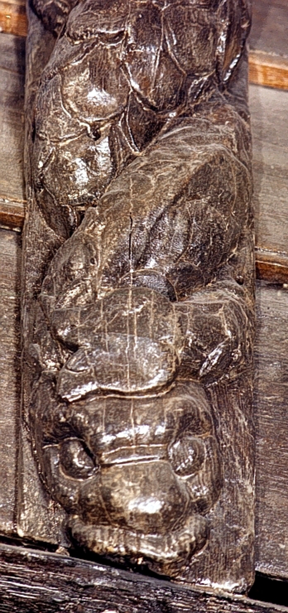Triton med mustascher och tjockt, vågigt hår. Armarna är reducerade till upprullade bladflikar. Runt midjan sitter ett band med en oval medaljong i mitten. Skulpturens fiskstjärt slingrar sig kring stjärten till en fisk eller delfin, vars huvud är synligt vid skulpturens fotända.



Bakstycket är kraftigt och snidat i ett med skulpturen. Baksidan är försedd med hak eller urtag. Skulpturens profil är svagt S-formad.

Skulpturen är kraftigt sliten och anletsdragen i det närmaste utplånade.



Text in English: Triton with moustache and thick wavy hair. The arms are replaced by rolled-up leafy decoration. Round the waist there is a girdle or belt with oval decoration. The fish tail is entwined with the tail of a fish or dolphin whose head can be seen at the foot of the sculpture.



The backboard is robust and carved in one piece with the figure. The back side of the backboard was carved with hollowed out grooves. The profile is slightly S-shaped.

The sculpture is badly worn, the features have been practically obliterated.