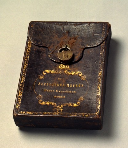 Klockarpostväska i form av ett låsbart (med hänglås) fodral. I fodralet förvaras en kvittensbok (PM 17200) från Simrishamns Pastors Expedition. På väskans framsida hänger i låsöglan ett litet hänglås i mässing och en till det tillhörande nyckel. Väskan som är i
läder har en i guldsnitt broderad kant och text enligt
signering/märkning inom en ornerad medaljong vilken överst kröns med kunglig krona, allt i guldsnitt.
