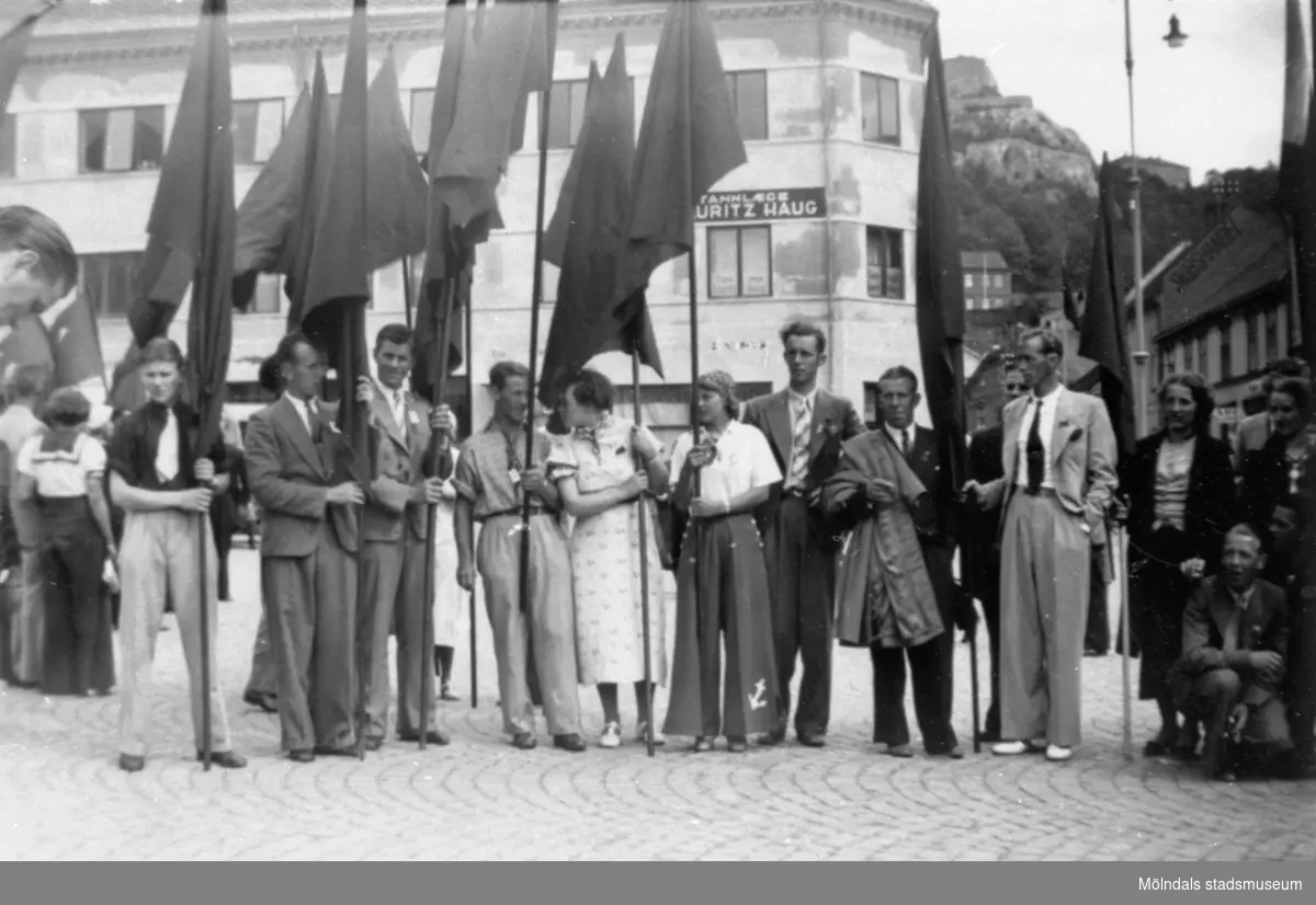 Flertalet män och kvinnor, medlemmar från SSU-föreningen i Mölndal. De står med flaggor, troligtvis politiskt möte, på torget i Halden. 1930-tal.