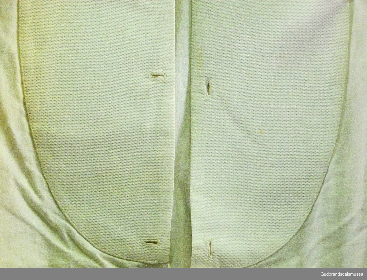 Hvit skjorte, mann, med stivet skjortebryst/bryststykke og brede kanter på ermene. Skjortebrystet har knappehull og åpning går helt ned. Tre hemper i front og en i nakke.