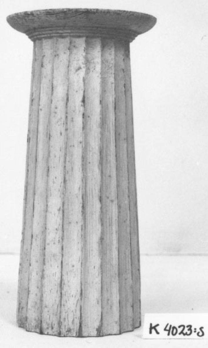 Pelare, eller kolonner, släta och räfflade (7 st halvkolonner, räfflade). 20 st. modeller av trä, avsedda som fasadprydnader. Pelarna är räfflade med halvcirkelformig basyta. De är vitmålade. Kapitälen är av närmast dorisk stil. Pelarna utgör modeller till gavelkonstruktion för nya inventariekammaren på varvet 1785-87. De sammanhängs troligen med en serie av gavelmodeller och pelare, som finns i kistan i sal 1 och vid norra delen av väggen i samma sal. (K 2244)

3/4 Kolonn, kannelerad, med kapitäl,Â målad i gråvitt.

H = 224mm  Bas D = 90mm  Kapitäl D = 110mm