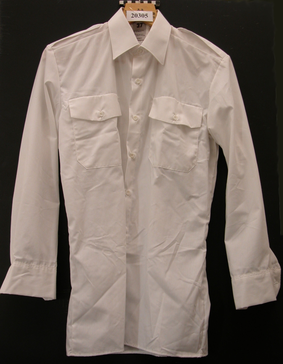 Skjorta, vit med lång ärm, 65% polyester, 35% bomull. Vita plastknappar.
Knäppning framtill, försedd med fast krage, fasta axelklaffar som knäpps med vita plastknappar. Påstickade bröstfickor med lock och knappar. Långa ärmar med enkal raka manchetter försedda med knappar. Insyningar i rygg som kan sprättas upp om skjortan behöver mer vidd.
Invändigt stämplad med Tre kronor och ?HEAB?. Lapp i nake ?37? samt skötselråd.
Storlek 37.