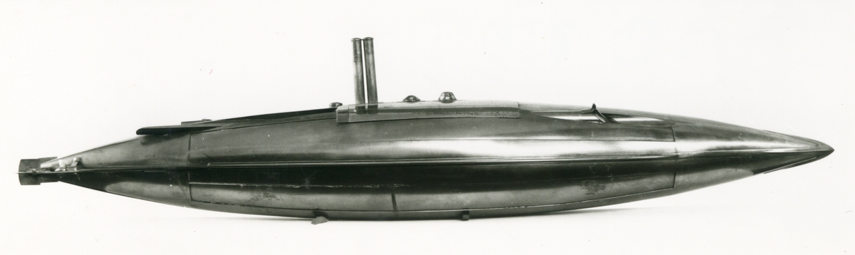 Spolformad. Stålplåt. Slingerköl, 2 propellrar av metall, 2 st höjd- och sidoroder parallellkopplade med gardjärn. 2 st höj- och sänkbara periskop av mässing. 4 st rännor för torpeder, därav 2 med luckor framtill, dels 2 för magasinering. Kort cylinder, rörlig i ränna midskepps akterut. Torpedrännorna täckas av långa stållock. Tillhörande 4 st torpeder av massiv mässing. Eneroths förslag till ubåt 1897. UndervattenstorpedFöremålets form: Spolformad.
