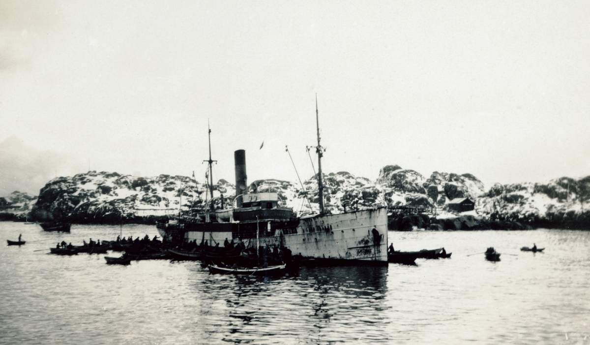 Agnbåten d/s "Rikard Kaarbø", omgitt av robåter, med bygning og fiskehjell i vinterlandskap i bakgrunnen.