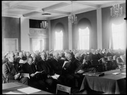 Gruppe menn sittende på stolrader med møtepapirer hendene.Sa