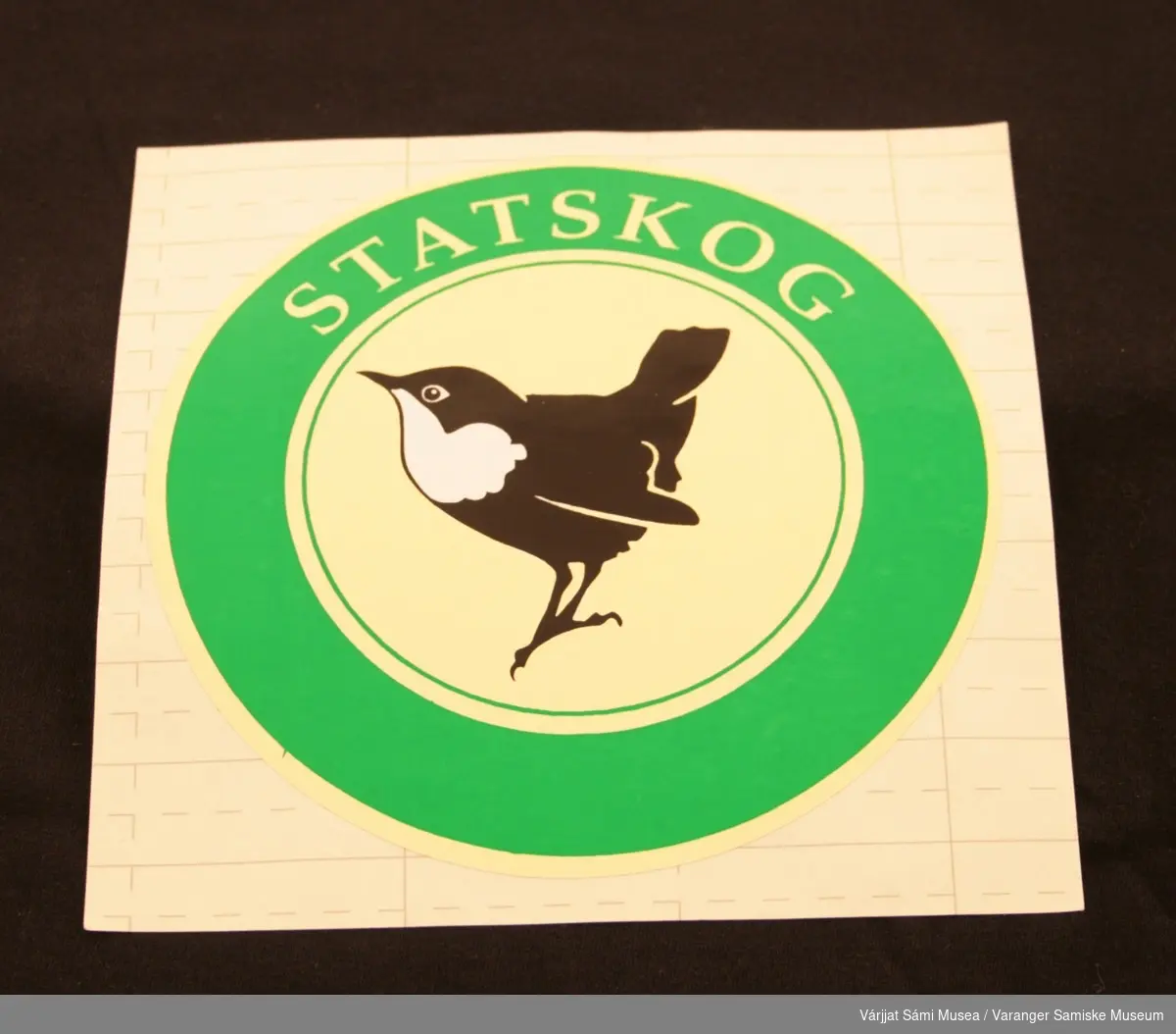 To runde klistremerker med Statskog logo: Fossekall i midten på lysegul bakgrunn, omgitt av en grønn sirkel med påskriften "Statskog" med lysegule bokstaver.
