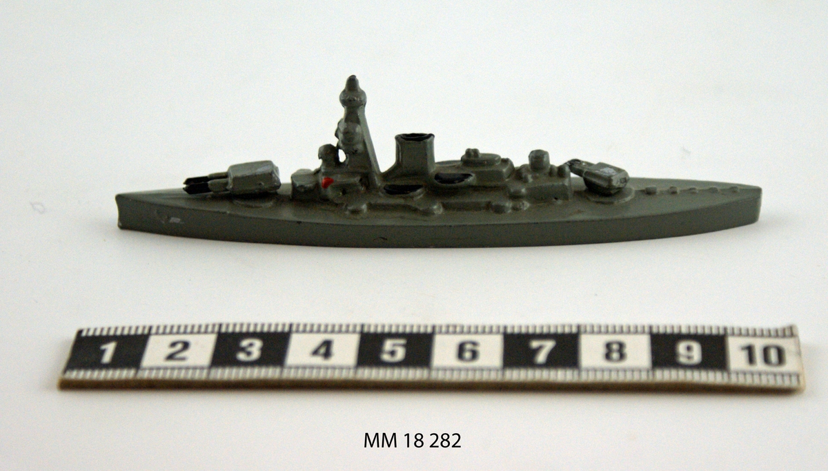 Fartygsmodell i form av pansarskeppet Gustav V gjuten av metall i ett stycke, målad i grått. Plan botten. Artilleripjäser i för och akter, dubbla vridbara. Mast och livbåtar med svart kapell, fyra stycken.