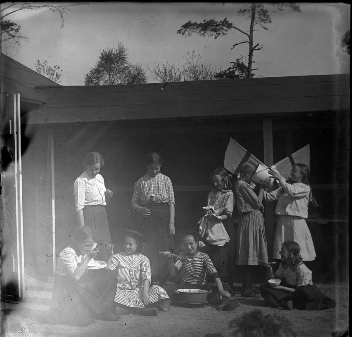 'Gruppbild, flickor, 1 kvinna (till vänster) och ev. 1 pojke, stående och sittande på marken framför byggnad. De är i färd med att äta. 1 flicka dricker från en hink. Glasflaskor. Tallrik. Slev. Gryta. Flaggor. Elever? ::  :: Ingår i serie med fotonr. 5415:1-10.'