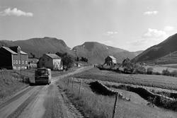 Buss på veien, hesjer til høyre og gårdsbebyggelse i bakgrun