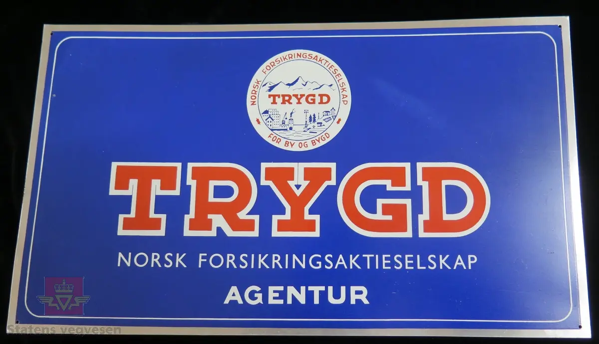 Flerfarget rekantgulært reklameskilt med teksten "TRYGD NORSK FORSIKRINGSAKTIESELSKAP AGENTUR". Skiltets bakside har ingen tekst.