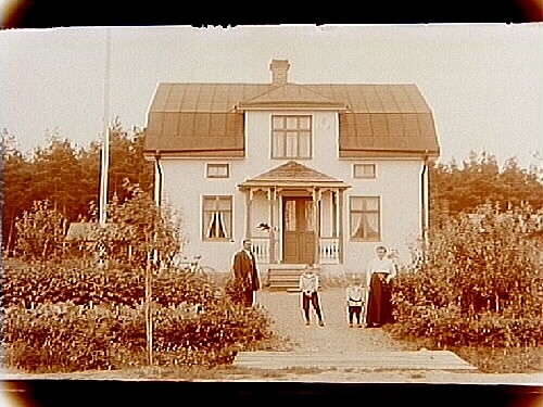 En och en halvvånings bostadshus med frontespis och veranda, familj 4 personer framför huset.
Axel Karlsson
