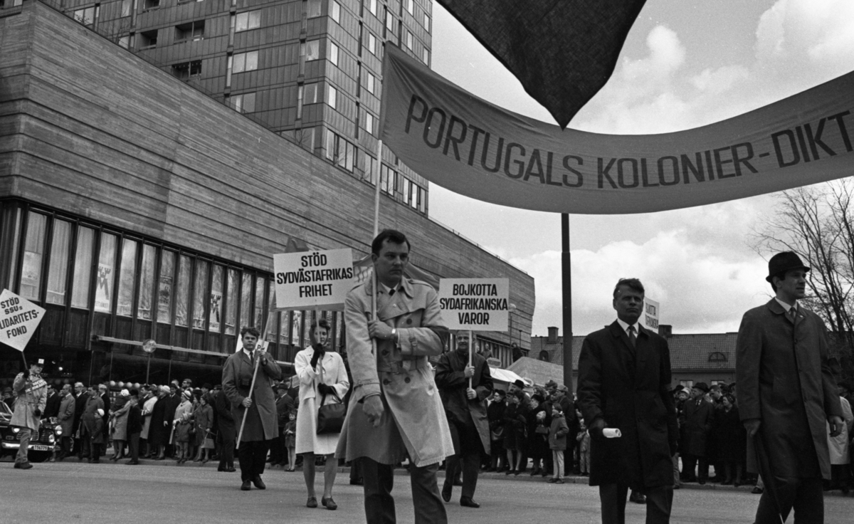 Första maj demonstration 2 maj 1967

En man klädd i vit rock kommer gående i ett första-majtåg utanför varuhuset Krämaren i Örebro med en banderoll med texten: "Portugals kolonier- diktatur." Han håller i en stång i ena hörnet av den stora banderollen. Två andra herrar går bredvid honom. Fler personer med skyltar kommer gående i bakgrunden.