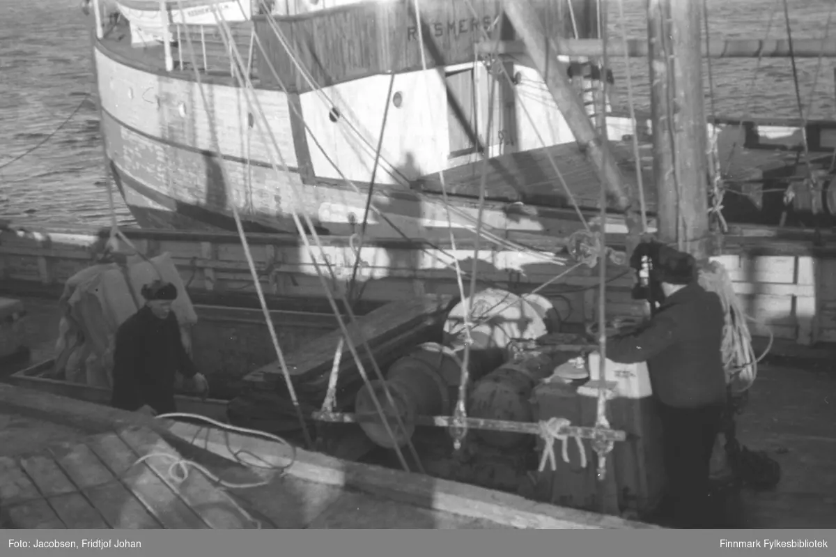 Lossing av forsyninger i Hammerfest vinteren 1945-46. To menn iført mørke klær og skinnluer står på dekket. En står i lasterommet og den andre betjener vinsjen. På trekaia ligger en lasteplatt med tau. En annen fraktebåt ligger fortøyd inntil båten nærmest kamera.