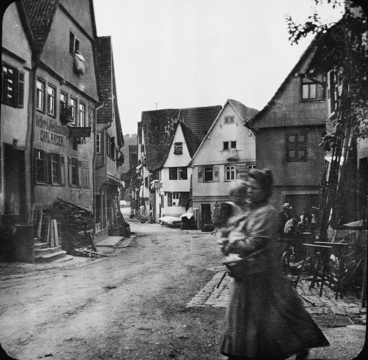 Skioptikonbild med motiv av kvinna med barn i famnen, på gata i Besigheim hörnet Kirchstrasse/Türkengasse.
Bilden har förvarats i kartong märkt: Resan 1907. Besigheim 10. 30